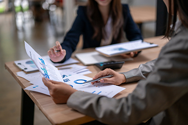 Businesswomen working together analyzing financial charts and documents at office desk Businesswomen analyzing financial charts and documents, discussing company performance and planning strategies