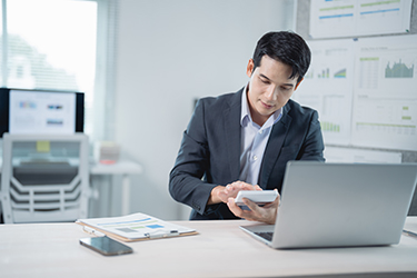Young businessman calculating and working with laptop and documents on desk in modern office, charts graphs diagrams and reports on whiteboard on background Young businessman calculating and working with laptop and documents on desk in modern office, charts graphs diagrams and reports on whiteboard on background