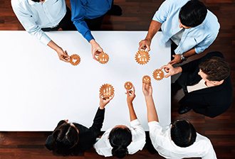 Collaborative Teamwork in a Business Setting with Diverse Professionals Engaging in Problem Solving Using Gears on a Table. Amity A diverse group of professionals collaborates around a table, exchanging ideas and holding gears to symbolize teamwork and innovative problem-solving in a business environment.